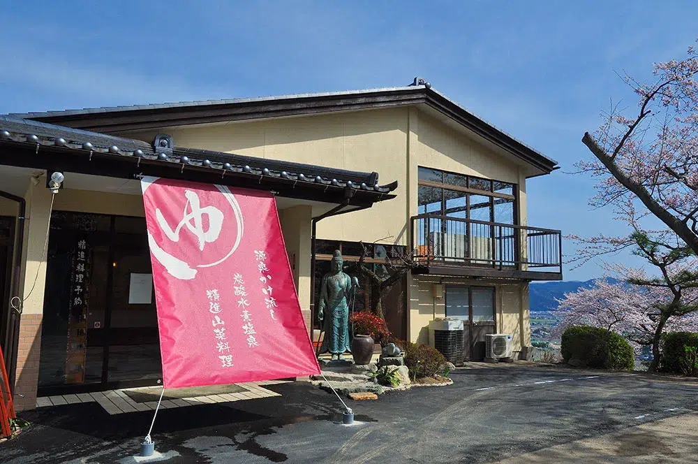 Yugenosato's entrance in cherry blossom season, with a red banner advertising its source-fed hot spring and shojin mountain vegetable cuisine
